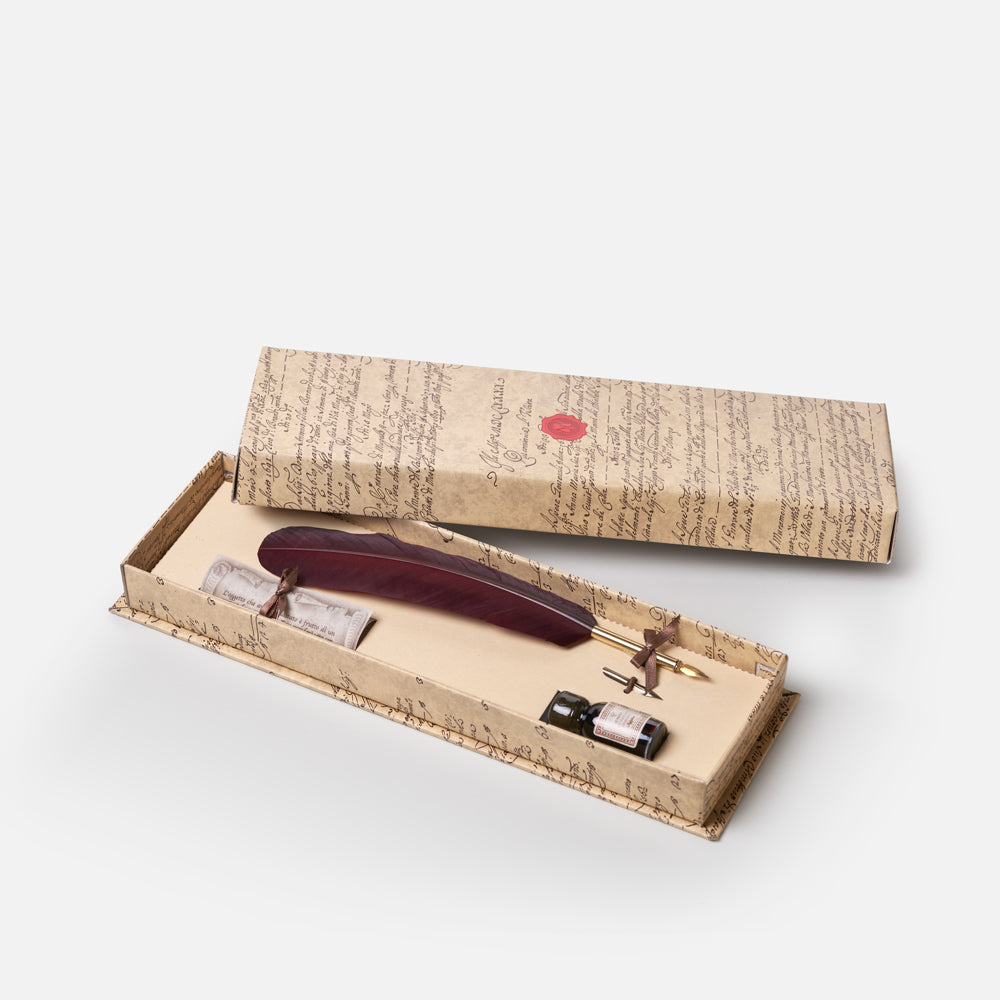 A vintage-style writing set displayed in an open decorative box with the lid placed behind it. The set includes a dark reddish-brown feather quill, a gold metal nib, small ink vials, and matching script-print packaging with a small red seal.